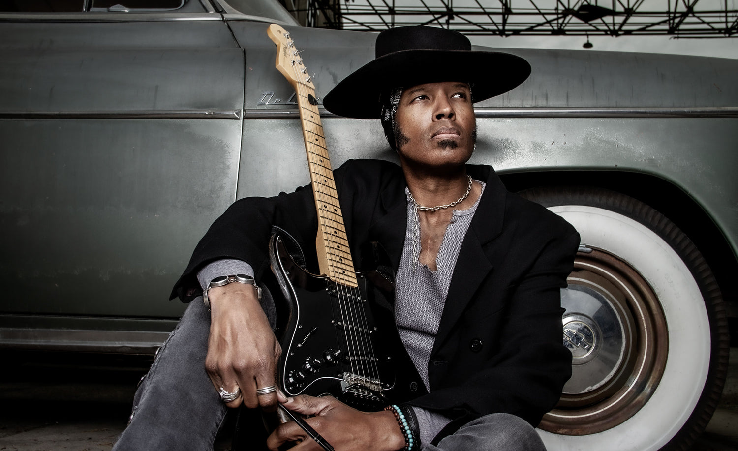 Male musician portrait Jeau James wearing black cowboy hat with black overcoat sitting down against old green car