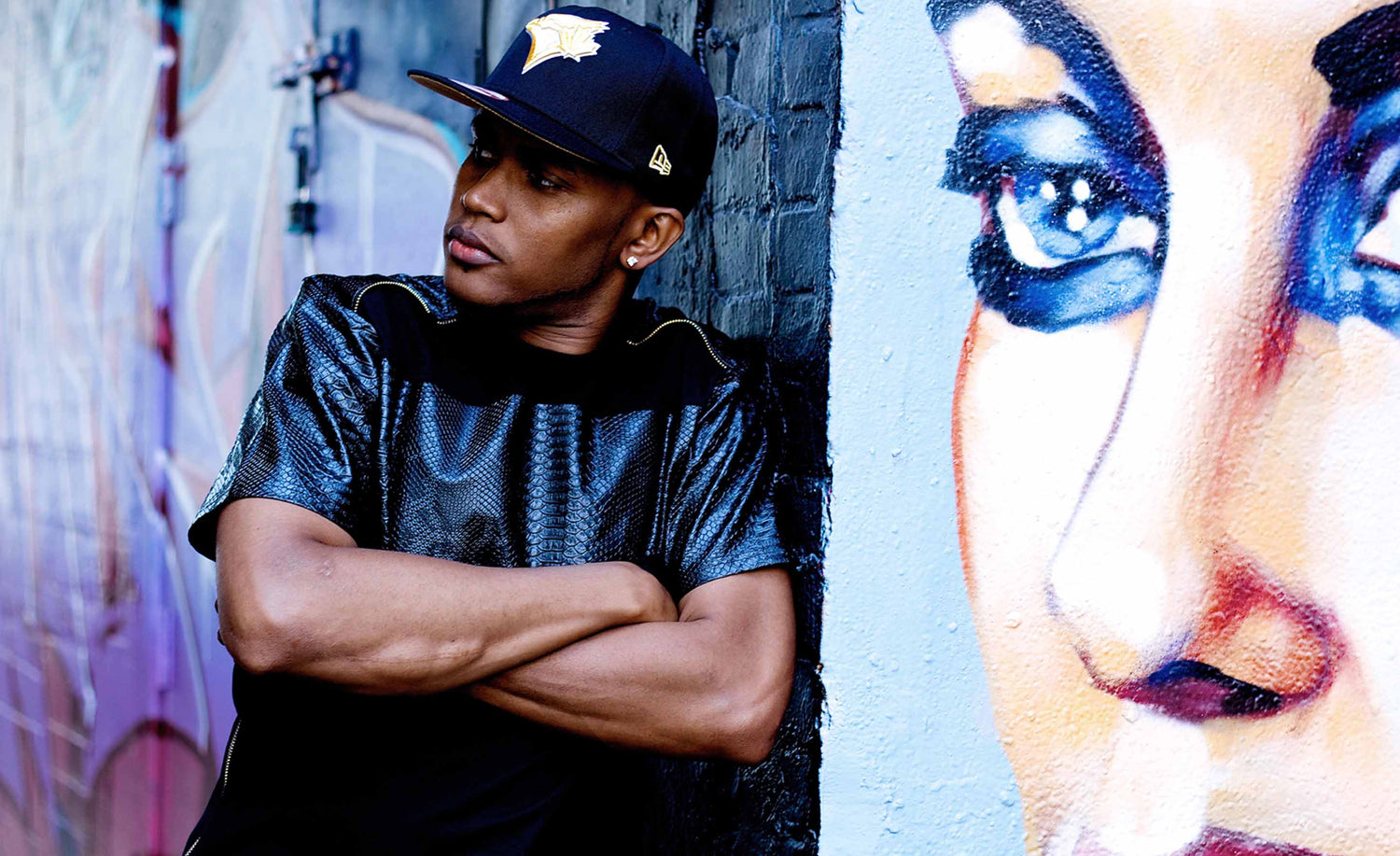 Musician portrait Fiyah MC leaning against black brick wall with graffiti of womans face beside him