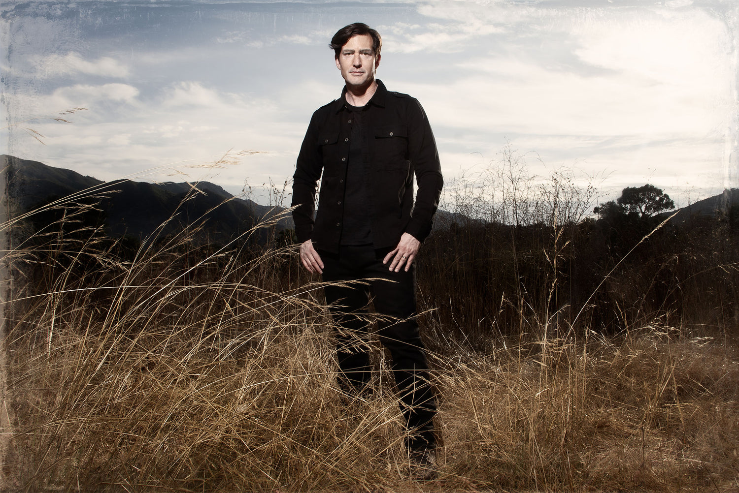 Composer Portrait Dino Meneghin standing in long golden grass field wearing black denim