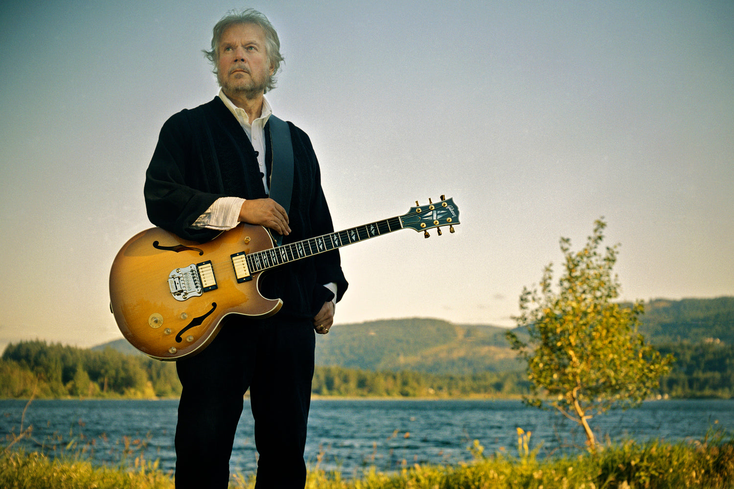 Randy Bachman portrait with guitar standing by lake looking off into distance