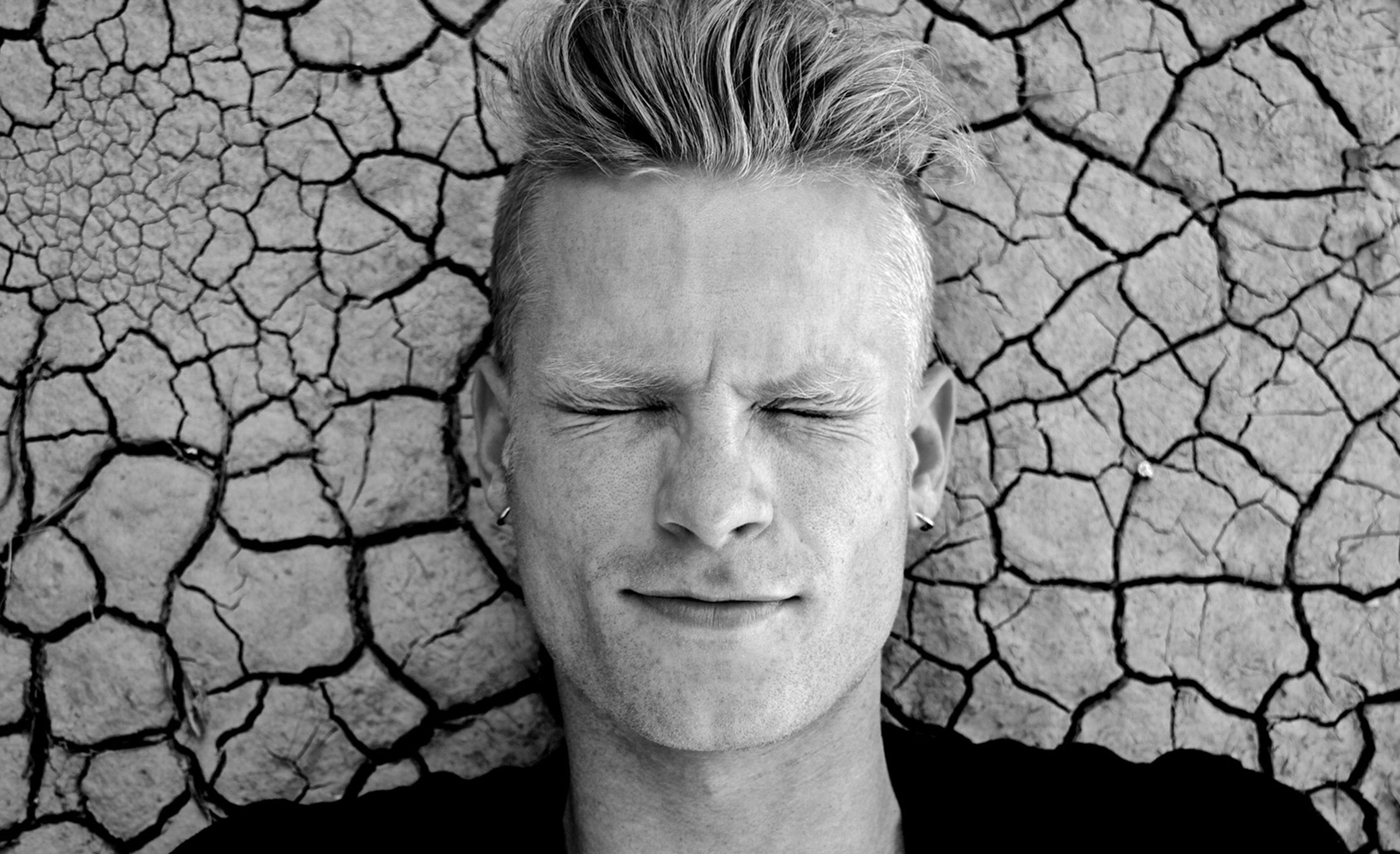 Black and white photo of mans face with eyes closed lying on cracked dry ground 