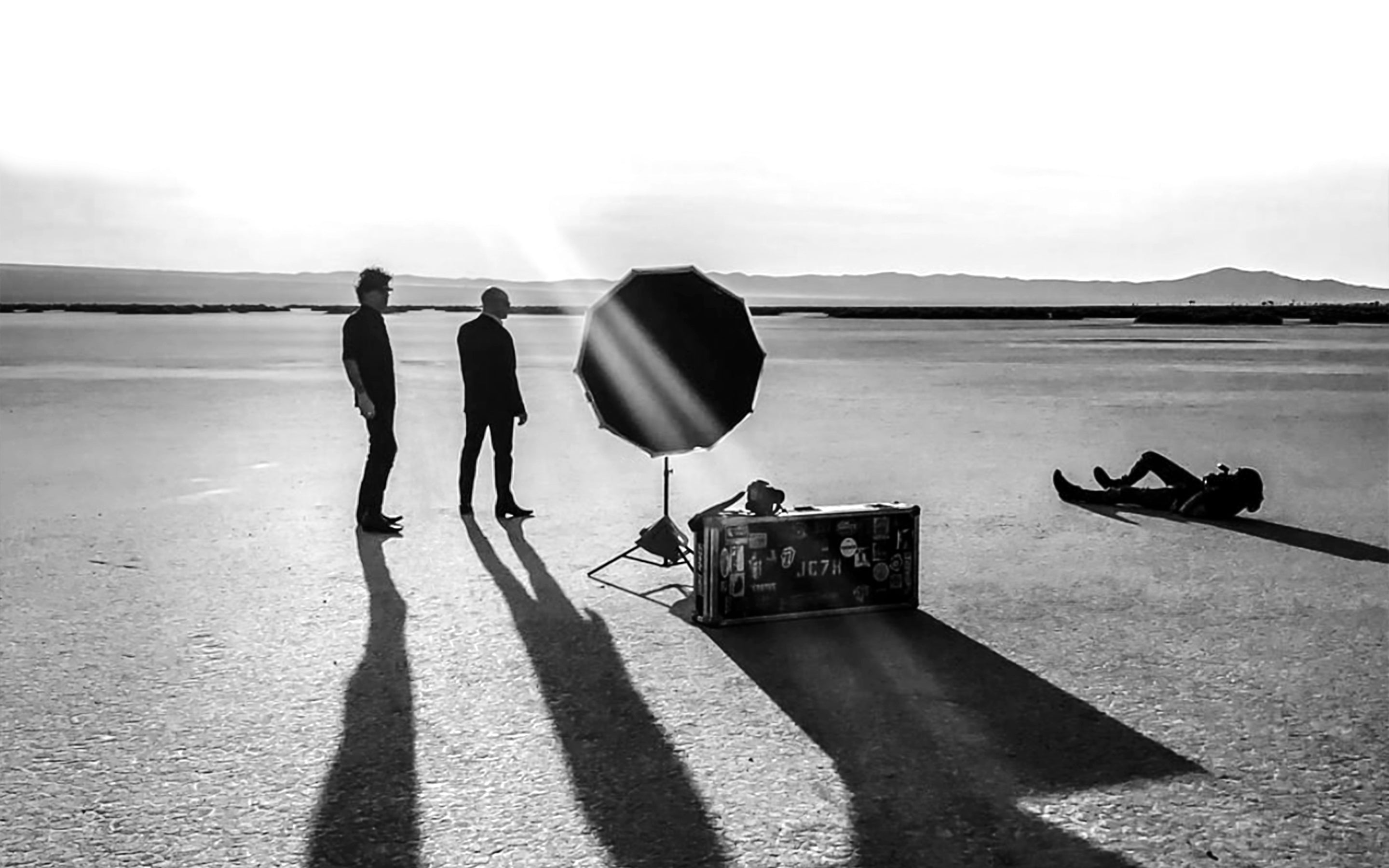 Black and white bts photo Mark Maryanovich lying on ground while photographing 7Horse standing in desert guitar and case beside reflector in front of them