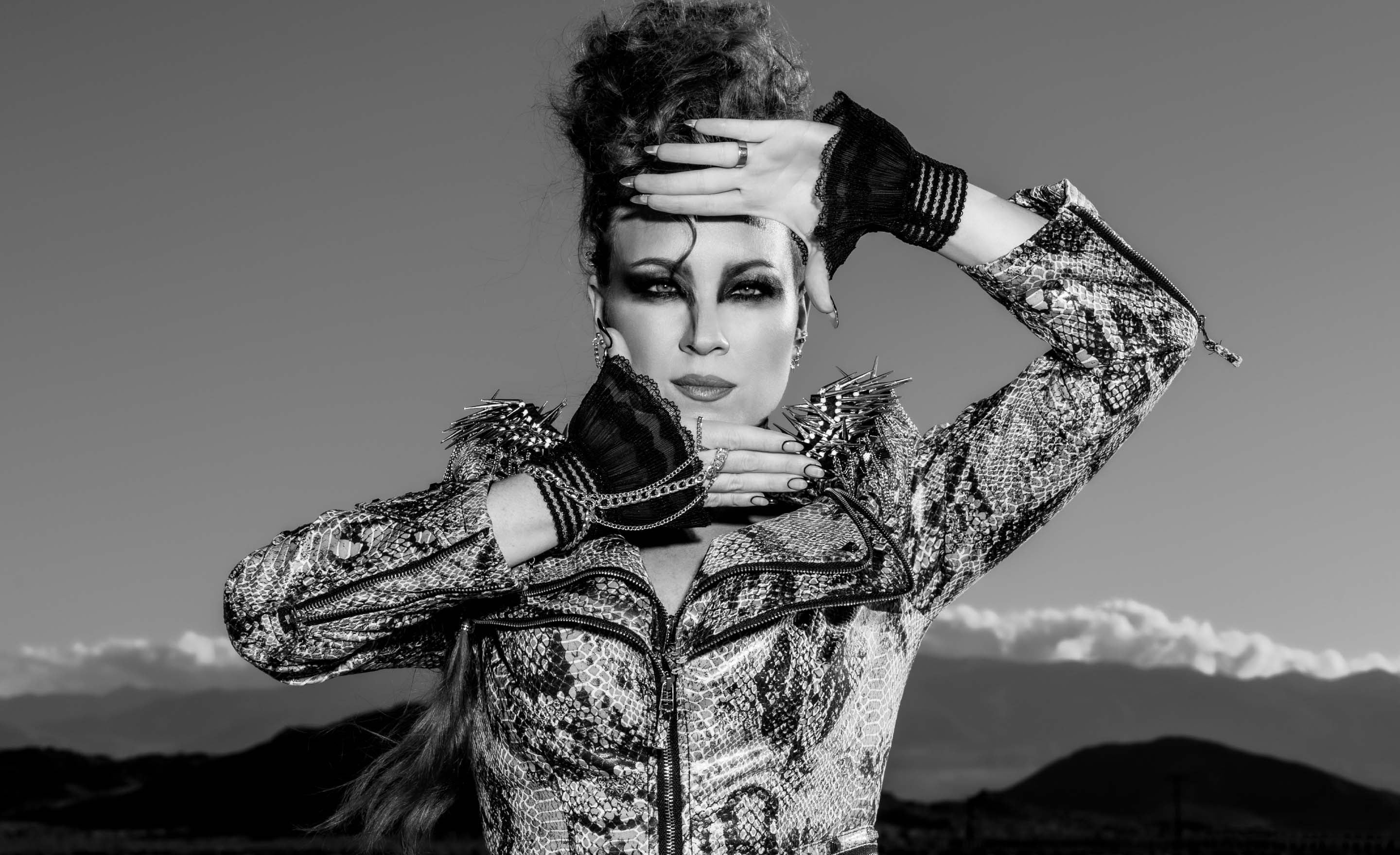 Black and white portrait musician Tarra Layne with hands framing face standing in front of mountain range with low clouds on the horizon behind her